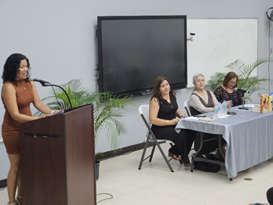 Las profesoras Emma Domenech Flores y Evelyn Jiménez Rivera junto a la escritora Esmeralda Santiago y la Dra. Rebeca Franquí como oradora parada ante un podio.