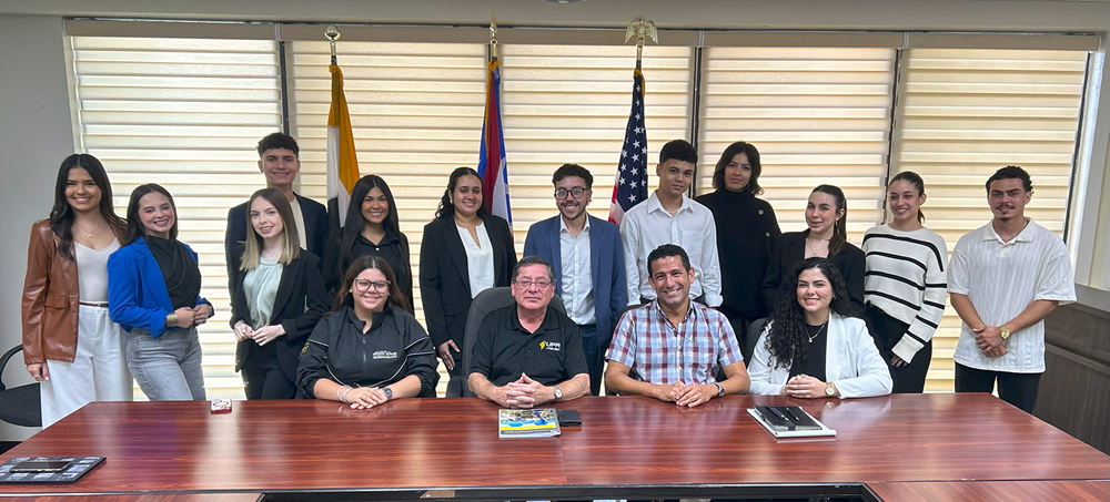 Fotografía grupal de estudiantes y personal de la UPR Arecibo reunidos en una sala de reuniones, posando frente a banderas institucionales y nacionales. Visten formal o semiformal y reflejan un ambiente profesional y de orgullo universitario.