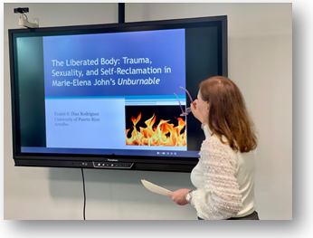 Ponente universitaria presenta en el Ciica Symposium sobre el libro Unburnable, con una diapositiva que trata temas de trauma, sexualidad y auto-reclamación. La imagen destaca un entorno académico con pantalla digital y proyección temática con fuego.