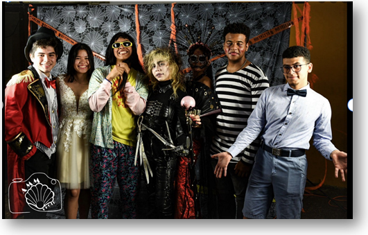 Grupo de siete personas disfrazadas de personajes como Edward Scissorhands, un maestro de ceremonias, un nerd y otros creativos atuendos. Posan con actitud alegre frente a un fondo decorado con telarañas y cintas naranjas, durante el evento Scary Con en UPRA.
