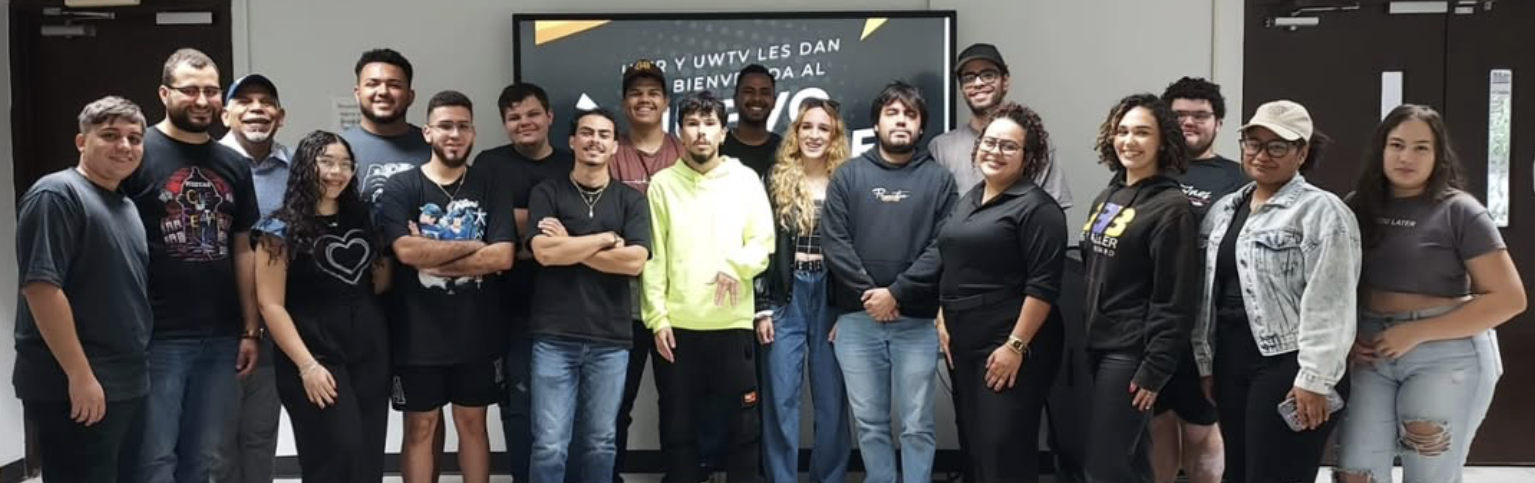 Grupo de 21 personas jóvenes posando en un pasillo frente a un cartel de bienvenida. La mayoría viste ropa casual y muestra expresiones amistosas. El fondo incluye dos puertas oscuras y un cartel central con texto parcialmente visible.