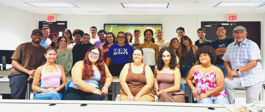 Fotografía grupal de estudiantes junto al profesor José Fonseca del Departamento de Comunicación Tele-Radial dentro de un salón de clases. Aproximadamente veinte estudiantes posan sonrientes frente a la cámara, distribuidos en dos filas, con cinco personas sentadas en la primera fila y las demás de pie detrás. Al fondo se ven una pantalla, puertas con letreros de «EXIT» y el mobiliario del salón.