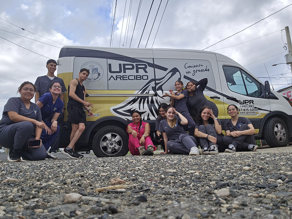 Grupo de estudiantes de tecnología veterinaria posa alegremente frente a una guagua rotulada de la UPR Arecibo. La escena, tomada desde el suelo, transmite espíritu de equipo en un entorno institucional.