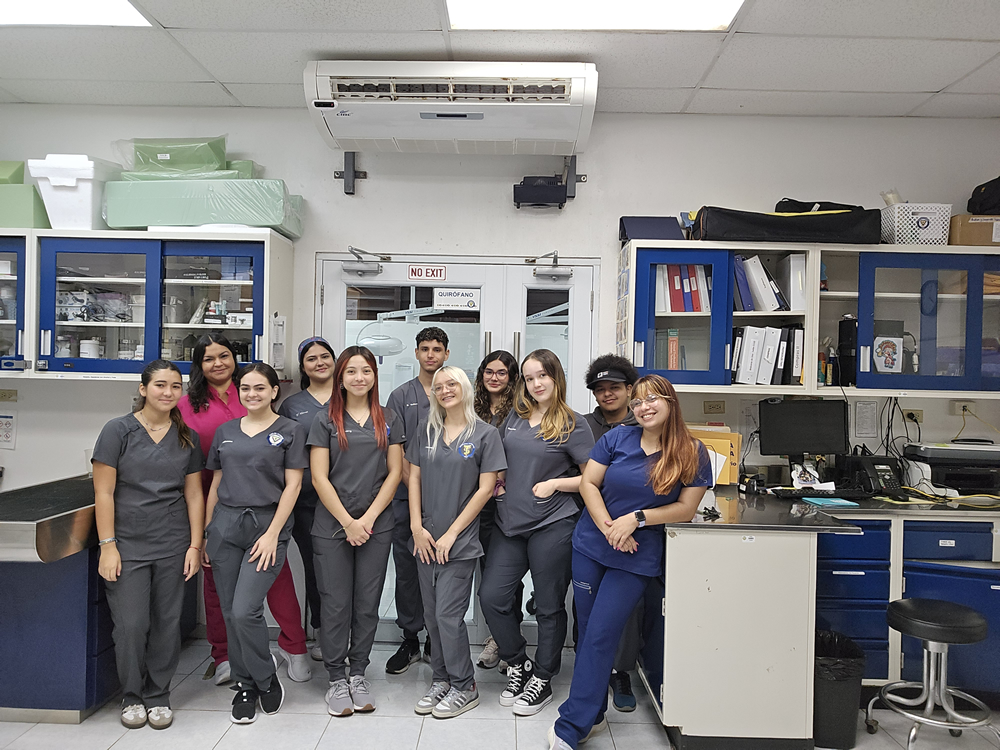 Estudiantes del Programa de Tecnología Veterinaria de la UPR en Arecibo posan dentro de un laboratorio clínico, usando uniformes médicos y rodeados de equipos, gabinetes y materiales de trabajo.