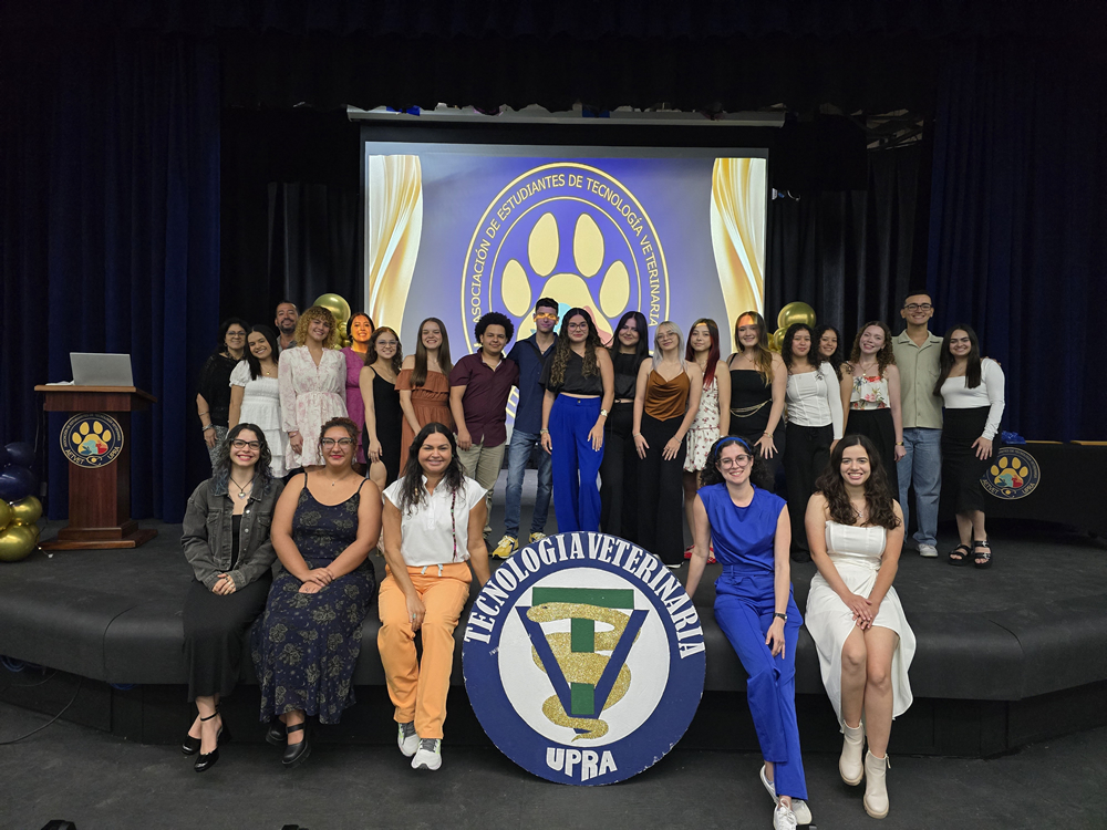 Grupo de estudiantes del Programa de Tecnología Veterinaria de la UPR en Arecibo posan en el Teatro decorado con emblemas institucionales y globos, durante una ceremonia o actividad especial. Al frente se exhibe un gran escudo con el nombre del programa, y al fondo se proyecta el logotipo de la Asociación de Estudiantes.