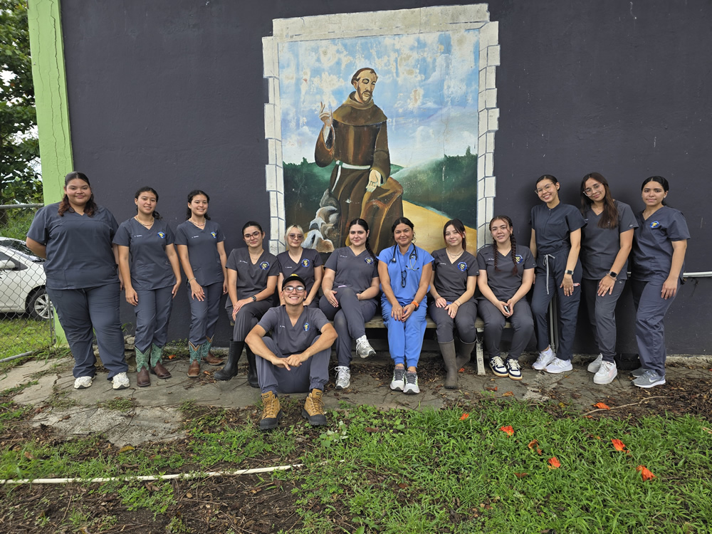 Grupo de estudiantes y la Dra. Rebeka Sanabria del Programa de Tecnología Veterinaria de la UPR en Arecibo posan frente a un mural de San Francisco de Asís. Visten uniformes clínicos y están al aire libre junto a una pared pintada y un área verde.