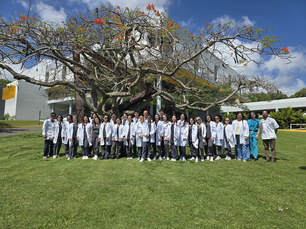 Estudiantes del Programa de Tecnología Veterinaria de la UPR en Arecibo posan al aire libre con batas blancas frente a un árbol frondoso y al edificio administrativo principal, en un ambiente académico soleado y natural.
