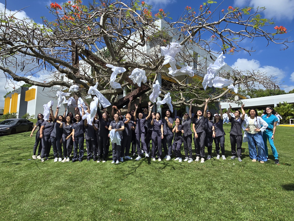 Estudiantes del Programa de Tecnología Veterinaria de la UPR en Arecibo celebran al aire libre, lanzando sus batas blancas sobre un árbol en un gesto simbólico de culminación académica. Están sonrientes, vestidos con uniformes clínicos oscuros, frente a un edificio moderno del campus, en un ambiente soleado y festivo.