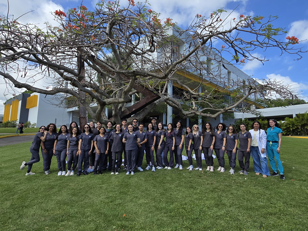 Estudiantes del Programa de Tecnología Veterinaria de la UPR en Arecibo posan organizadamente frente a un árbol emblemático del campus tras una celebración. Visten uniformes clínicos oscuros, y se les ve sonrientes junto a la Dra. Rebeka Sanabria y la Técnica de Laboratorio. El fondo muestra el edificio universitario bajo un cielo soleado.