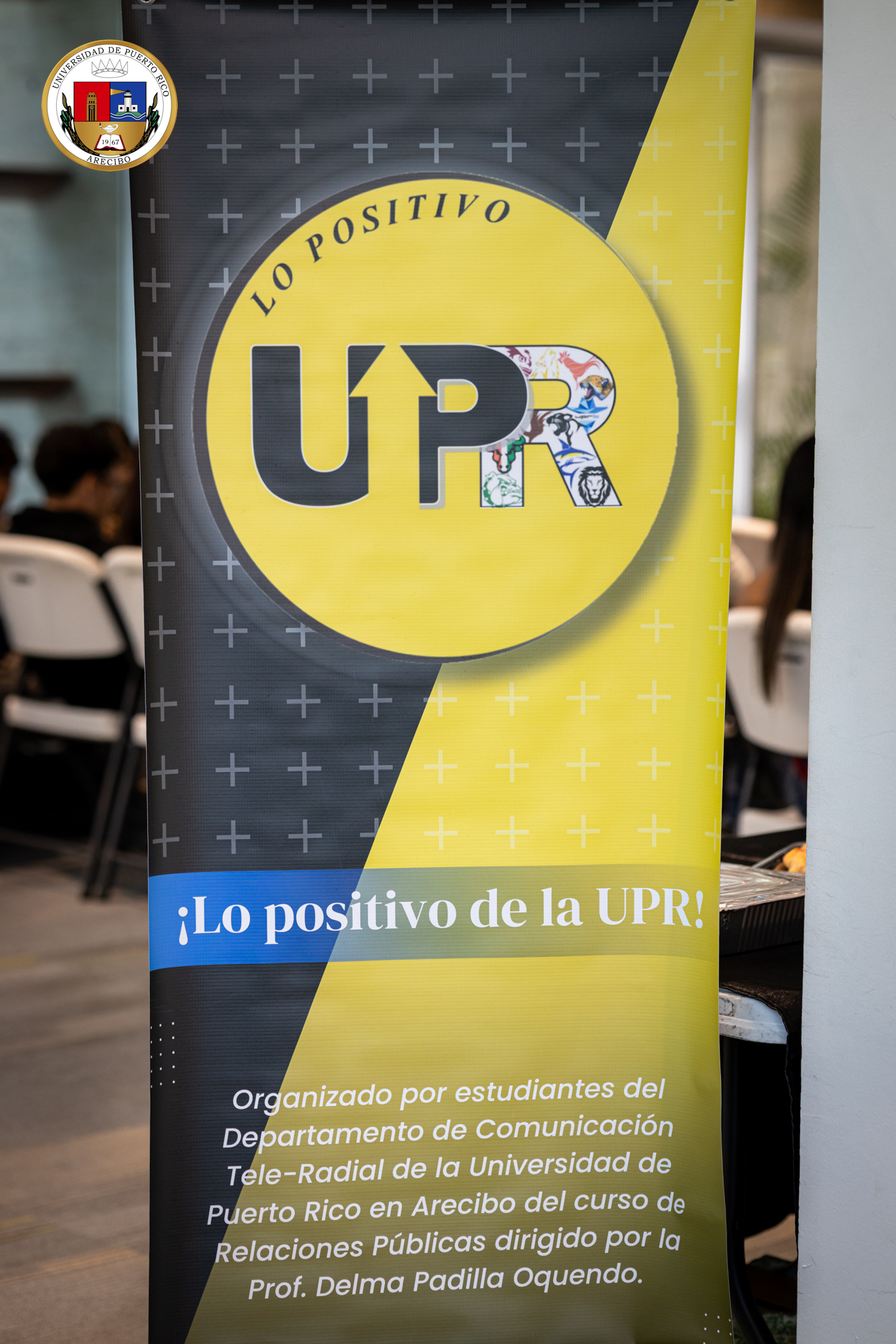 2K2A3906 Banner vertical de la UPR con fondo en tonos amarillo y negro, el eslogan “Lo positivo de la UPR” y un texto informativo sobre un evento organizado por estudiantes del Departamento de Comunicación Tele-Radial en UPR Arecibo.