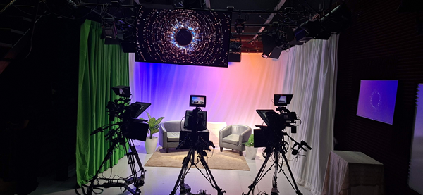 Dept-COMU Estudio de televisión del Departamento de Comunicación Tele-Radial de la UPR en Arecibo, equipado con tres cámaras profesionales sobre trípodes, iluminación escénica y pantallas. En el área central hay dos sillones grises preparados para entrevistas, con un fondo iluminado en tonos azul, violeta y anaranjado. A la izquierda se observa una cortina verde para chroma key y, en la parte superior, una pantalla con un gráfico circular luminoso.