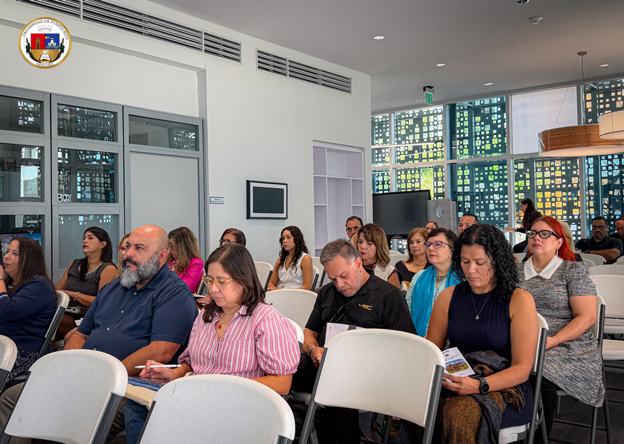 Fotografía de los asistentes a la Actividad Gerencial número 24 de la Universidad de Puerto Rico en Arecibo. Varias filas de personas adultas están sentadas en sillas blancas dentro de un salón moderno con grandes ventanales. Algunos participantes escuchan atentamente, mientras otros escriben en cuadernos o leen folletos. En primer plano destacan un hombre calvo con barba canosa, una mujer con blusa de rayas rosadas y blancas que toma notas, y una mujer de cabello rizado que consulta un folleto. El escudo de la Universidad de Puerto Rico aparece en la esquina superior izquierda.