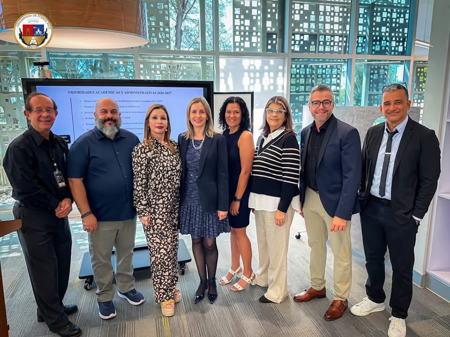 Fotografía de ocho integrantes de la Universidad de Puerto Rico en Arecibo, seis mujeres y dos hombres, posando sonrientes frente a una pantalla con la presentación «Prioridades académicas y administrativas 2026-2027». La actividad se desarrolla en un espacio de oficinas iluminado por grandes ventanales y corresponde a la Actividad Gerencial número 24 de la institución.